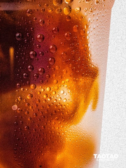 This is a photorealistic close-up image capturing the surface of a glass filled with a dark amber liquid, likely iced tea or cola, covered in condensation. The visual aesthetic is hyper-detailed and textural, focusing on the interplay of light and water droplets. The color language is dominated by a warm, rich palette of deep amber, burnt orange, and dark brown tones within the liquid, contrasted against the cool, translucent white of the condensation and a bright, overexposed white background on the right. Saturation is high within the liquid's hues, creating a vibrant, glowing effect, while contrast is strong between the dark liquid and the bright highlights on the droplets. The lighting approach appears to be backlit or side-lit from the right, causing the liquid to glow with an internal luminescence and creating sharp, specular highlights on each individual water droplet, enhancing their three-dimensional form. The texture and material feel are intensely tactile; the glass surface is obscured by a dense layer of condensation, with droplets ranging from tiny, mist-like beads to larger, globular drops that catch and refract the light, creating a wet, cold, and refreshing impression. Compositionally, the image is a tight, cropped shot that fills the frame with texture, using a shallow depth of field where the droplets on the near surface are in sharp focus while the liquid and background blur, emphasizing the macro details. The shape language is entirely organic, defined by the irregular, spherical forms of the water droplets and the fluid, amorphous shapes of the liquid and ice within, with soft edges on the blurred elements and crisp, hard edges on the focused droplets. The mood and atmosphere evoke a sense of refreshment, coolness, and sensory immediacy, with a high-energy visual weight concentrated on the intricate, glittering surface details.