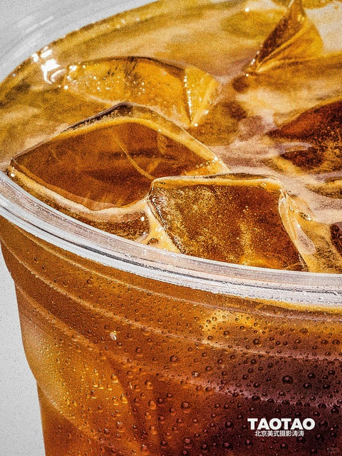 This image presents a hyper-realistic, close-up photographic rendering of a cold beverage in a clear plastic cup, characterized by a highly textured and tactile aesthetic. The visual style is photorealistic with a focus on material detail, capturing the intricate interplay of light and liquid. The color palette is dominated by warm, saturated amber and deep caramel tones from the beverage, contrasted against the cool, translucent white of the ice and the neutral gray background, creating a strong warm-cool dynamic. Lighting is directional and hard, coming from the upper left, casting sharp, defined highlights on the ice cubes and the rim of the cup while creating deep, rich shadows within the liquid, enhancing the sense of volume and chill. The texture and material rendering are exceptionally detailed, emphasizing the condensation droplets beading on the cup's exterior, the porous, crystalline surface of the ice, and the effervescent bubbles suspended in the dark liquid, creating a palpable sense of coldness and refreshment. The composition is a tight, angled close-up that crops the top of the cup, focusing intensely on the surface of the drink and the ice, with a shallow depth of field that keeps the foreground sharp and slightly softens the background elements. The shape language is a mix of organic and geometric forms: the organic, irregular shapes of the melting ice contrast with the precise, manufactured curve of the cup's rim and the circular pattern of condensation droplets. The overall mood is one of intense refreshment, tactile appeal, and summery coolness, with a high-energy visual weight derived from the sharp contrasts and detailed textures.