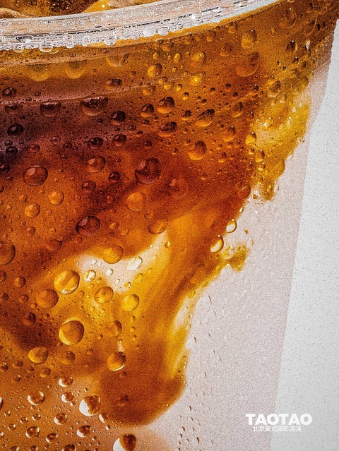This is a hyper-detailed, photorealistic close-up photograph of a carbonated amber beverage, likely iced tea or a dark soda, contained within a frosted glass. The visual aesthetic is intensely textural and macro-focused, emphasizing the physical properties of the liquid and container. The color language is dominated by a warm, rich spectrum of amber, caramel, and deep brown tones within the liquid, contrasted against the cool, desaturated off-white of the frosted glass. Saturation is high within the liquid itself, with vibrant golden highlights where light passes through, while the glass area is muted. Contrast is strong, with deep shadows within the liquid's depth and bright, specular highlights on the bubbles and glass surface. The lighting approach appears to be directional from the upper left, creating a luminous glow through the translucent liquid and casting soft shadows that enhance the three-dimensionality of the bubbles. The quality is hard enough to create sharp reflections but diffused by the liquid and glass, resulting in a glowing, backlit effect. Texture and material feel are paramount: the liquid is rendered with a viscous, effervescent quality, filled with countless bubbles of varying sizes that appear spherical and three-dimensional. The glass has a distinct frosted, condensation-covered texture, with tiny water droplets and a grainy, matte finish that contrasts with the smooth, glossy bubbles. Compositionally, this is an extreme close-up with a diagonal framing that cuts across the glass edge, creating a dynamic division between the dense, textured liquid and the simpler frosted surface. Depth is shallow, with the focus sharp on the bubbles and glass texture in the mid-ground, while the background liquid blurs slightly. Shape language is overwhelmingly organic, defined by the irregular, spherical forms of the bubbles and the fluid, amorphous shapes of the liquid, with no hard geometric lines. The edge treatment is soft where the liquid meets the glass, with a blended, seeping quality. The mood and atmosphere are refreshing, tactile, and immersive, conveying a sense of chill and effervescence through the visual weight of the condensation and the energetic, upward movement implied by the bubbles.