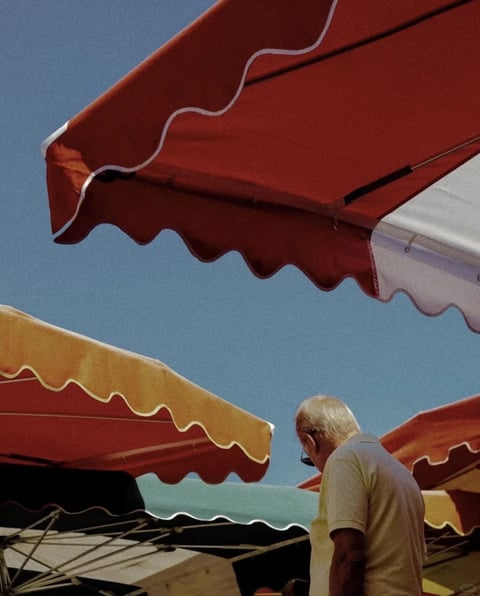 The image exhibits a photorealistic aesthetic with a distinct film grain texture, suggesting a vintage or analog photographic quality. The color language is defined by a high-contrast, saturated palette dominated by warm, sun-drenched tones: deep terracotta reds, golden yellows, and crisp whites, all set against a clear, vibrant cerulean blue sky. The lighting is harsh and direct, characteristic of bright midday sun, creating sharp, well-defined shadows and bright, almost blown-out highlights on the awnings and the subject's white hair. Texture and material feel are rendered with tactile clarity; the canvas of the awnings appears thick and slightly stiff, with visible stitching and scalloped edges, while the man's polo shirt shows soft, woven fabric folds. Compositionally, the frame is densely layered with overlapping diagonal planes created by the awnings, which slice across the image and create a sense of compressed depth, with the man positioned in the lower right quadrant, partially obscured. Shape language is a mix of the rigid, geometric lines of the awning frames and the soft, organic curves of their scalloped edges and the human figure. The overall mood is one of quiet, sun-baked stillness and leisurely observation, evoking a warm, nostalgic atmosphere of a Mediterranean summer day.
