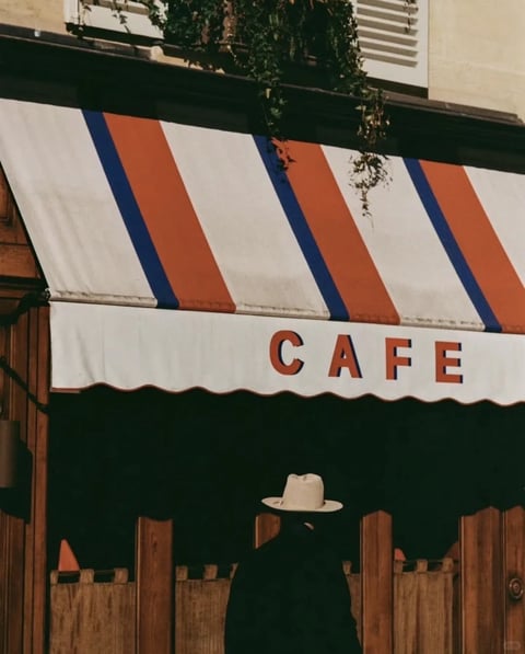 The image presents a photorealistic, film-like aesthetic with a warm, nostalgic color palette dominated by terracotta orange, cream white, and deep navy blue, accented by rich wood tones and deep, shadowed greens. The lighting is soft and diffused, suggesting an overcast day or shaded environment, casting gentle, indistinct shadows that contribute to a calm, contemplative mood. Textures are rendered with subtle realism: the canvas awning shows slight wear and fabric weave, the wooden fence and facade display grain and matte finishes, and the foliage above has a soft, organic detail. Compositionally, the frame is tightly cropped, focusing on the awning and the solitary figure below, creating a sense of intimate, almost voyeuristic depth through layered planes of the awning, fence, and shadowed interior. Shapes are a mix of strong geometric lines from the awning stripes and fence posts, contrasted with the organic curves of the hanging plants and the soft silhouette of the hat. The overall atmosphere is one of quiet, cinematic stillness, evoking a vintage European street scene with a touch of melancholic elegance.