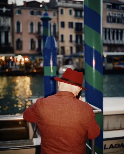 The image presents a photorealistic, cinematic composition with a shallow depth of field, focusing on the back of an older man in a textured rust-red blazer and a matching red fedora with a dark band. He is positioned in the foreground, leaning against a railing, creating a sense of intimate observation. The background is softly blurred, revealing a canal scene with historic, pastel-colored buildings (soft pinks, ochres, and creams) and distinctive blue-and-green striped mooring poles, suggesting a location like Venice. The color palette is warm and saturated, dominated by the man's rich red attire against the cooler, muted tones of the water and architecture, with golden bokeh highlights reflecting on the water's surface from distant lights. The lighting is soft and diffused, likely from an overcast sky or dusk, casting gentle shadows and enhancing the textures of the fabric and the weathered surfaces. The composition uses the vertical lines of the poles to frame the subject, creating a layered, three-dimensional feel with a clear foreground, middle ground, and background. The overall mood is contemplative and nostalgic, with a quiet, elegant energy conveyed through the subject's posture and the serene, blurred environment.