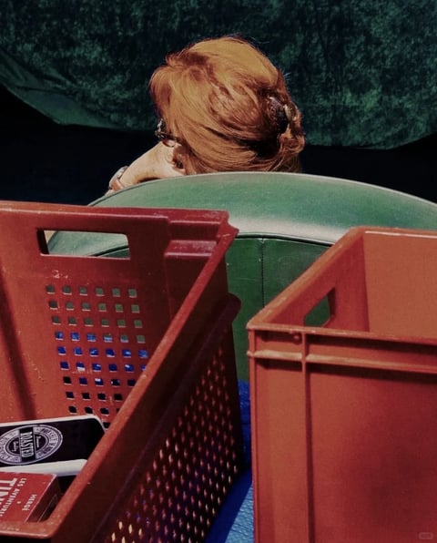The image exhibits a cinematic, filmic aesthetic with a soft, grainy texture reminiscent of analog photography, rendered with a shallow depth of field that keeps the foreground crates in sharp focus while the background figure and chair are softly blurred. The color language is dominated by a warm, earthy palette featuring deep rust-reds, muted terracotta, and olive greens, with subtle cool blue accents visible through the crate perforations, all rendered with moderate saturation and a slightly desaturated, vintage tonal quality. Lighting appears soft and diffused, likely from an ambient source, creating gentle gradients and soft shadows that emphasize the rounded forms of the chair and the hair's volume without harsh contrast. Textures are tactile and material-focused: the plastic crates show a matte, slightly worn surface with visible molding lines and perforations, the chair presents a smooth, supple leather-like appearance with visible stitching, and the hair displays fine, individual strands with a soft, glossy sheen. Compositionally, the frame is tightly cropped with a layered, overlapping arrangement where the geometric, angular crates in the foreground create a structured frame that contrasts with the organic, rounded forms of the chair and the person's head behind them, establishing a clear foreground-background separation. Shape language contrasts the rigid, rectilinear geometry of the crates with the soft, organic curves of the hair and chair, with clean edges on the manufactured objects versus softer, blurred edges on the human element. The mood is intimate, quiet, and slightly voyeuristic, conveying a sense of private contemplation or rest within a mundane, everyday setting, with a low-energy, visually weighted atmosphere focused on texture and form.