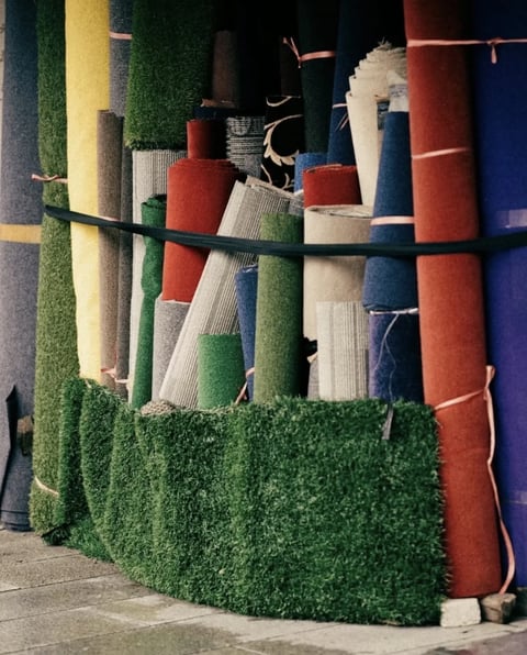 The image presents a photorealistic, tightly framed composition of rolled and stacked textile materials, likely carpets or rugs, arranged vertically against a wall. The visual aesthetic is grounded in documentary realism, capturing the tangible, tactile quality of the materials with sharp detail. The color language is rich and varied, dominated by deep, saturated earth tones—rust red, olive green, and mustard yellow—punctuated by cooler accents of slate blue and charcoal gray, all rendered under a soft, diffused, neutral-toned light that suggests an overcast day or shaded outdoor setting, minimizing harsh shadows. The lighting approach is even and flat, emphasizing the material surfaces without dramatic contrast. Texture and material feel are central, with the image highlighting the coarse, fibrous weave of the carpets, the smooth, taut surfaces of the rolled fabrics, and the shaggy, grass-like pile of the artificial turf in the foreground, creating a strong tactile impression. The composition is dense and layered, with the vertical rolls creating a rhythmic, almost architectural pattern that fills the frame, while the horizontal black strap and the curved base of the artificial turf provide structural counterpoints. Shape language is predominantly geometric and cylindrical from the rolls, contrasted by the organic, irregular texture of the turf. The overall mood and atmosphere are utilitarian and grounded, evoking a sense of craftsmanship, inventory, and the raw materiality of textiles in a market or workshop setting.