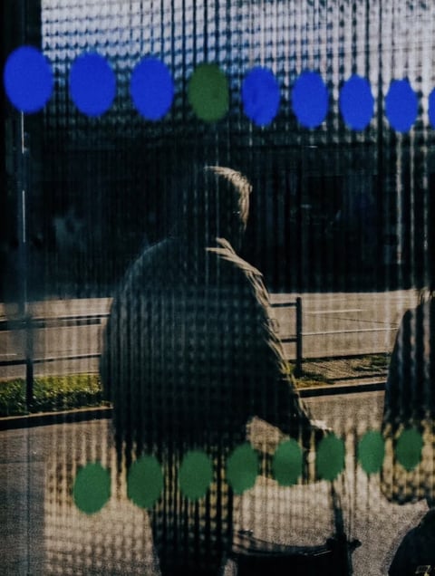 The image presents a moody, atmospheric street scene viewed through a semi-transparent, patterned surface, likely a window with a fine mesh or screen overlay that creates a consistent vertical line texture and a subtle grid-like distortion across the entire frame. The visual aesthetic is photorealistic yet heavily stylized by this textural filter, giving it a cinematic, slightly degraded film quality. The color language is dominated by deep, desaturated cool tones—charcoal blacks, slate grays, and muted blues—with selective, vibrant accents of electric blue and forest green from the out-of-focus bokeh circles in the upper and lower foreground. The lighting is low-key and directional, appearing to come from the upper left, casting strong, hard-edged shadows and creating high contrast between the silhouetted figure and the brighter, washed-out background. The texture and material feel are defined by the prominent screen overlay, which imposes a tactile, almost digital or woven grain over the scene, while the underlying elements—the person's coat, the wet pavement—suggest a damp, cool environment. Compositionally, the image uses a shallow depth of field, with the foreground bokeh and screen texture framing a central, silhouetted figure walking away, creating a sense of depth and voyeuristic distance. The shape language is a mix of organic forms (the human silhouette, the soft bokeh orbs) and strict geometric lines (the vertical screen pattern, the horizontal fence rails). The overall mood is introspective, somber, and slightly mysterious, evoking a feeling of urban isolation or a fleeting moment observed through a barrier.