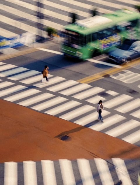The image presents a high-angle, slightly blurred urban street scene captured in a photographic style with a tilt-shift or miniature effect, creating a soft, dreamlike quality. The color language is defined by a warm, sun-drenched palette, with the asphalt rendered in cool grey-blue tones contrasted by the stark white of the crosswalk stripes and the warm, earthy terracotta of the pedestrian island. A vibrant green bus provides a strong color accent, its saturation slightly muted by the overall soft focus. The lighting is directional, casting long, sharp shadows from the pedestrians and the bus, indicating a low sun angle, likely late afternoon or early morning, which enhances the scene's depth and three-dimensionality. The texture is smooth and slightly diffused, with the asphalt, painted lines, and vehicle surfaces appearing clean and graphic, lacking granular detail due to the intentional blur. Compositionally, the image is structured by strong diagonal lines from the crosswalks and road markings, creating dynamic movement, while the high vantage point flattens the perspective, emphasizing the geometric patterns of the urban grid. The shape language is predominantly geometric, dominated by the parallel white rectangles of the crosswalks and the angular forms of the road and vehicles, with the organic shapes of the pedestrians providing scale and focal points. The overall mood is one of quiet, cinematic urban observation, evoking a sense of fleeting motion and sunlit solitude within a structured cityscape.
