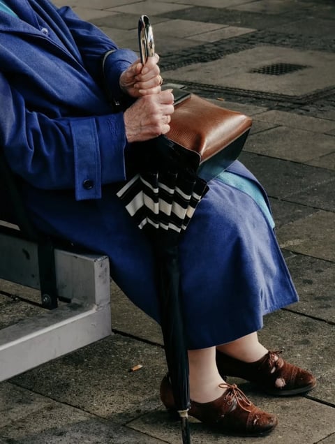 This is a photorealistic, high-contrast photographic image with a shallow depth of field, focusing on the lower half of an elderly person seated on a weathered, grey concrete bench. The composition is tightly framed from a slightly high angle, emphasizing the subject's lap, hands, and feet against a textured, grey stone pavement background. The dominant color palette is a rich, saturated cobalt blue from the subject's long coat and skirt, contrasted sharply with the warm, earthy tones of a brown leather handbag and matching brown suede lace-up shoes. The lighting is soft and diffused, suggesting an overcast day, with gentle shadows that define the folds of the fabric and the textures of the materials. The image highlights a variety of tactile surfaces: the heavy, matte wool of the coat, the smooth, polished leather of the bag, the soft, napped suede of the shoes, and the coarse, gritty texture of the stone ground. The subject's aged hands, with visible veins and wrinkles, firmly grip a closed black umbrella with a polished silver handle, which rests vertically between their legs. The umbrella features a distinctive black and white striped pattern near the handle. The overall mood is quiet, contemplative, and slightly melancholic, with a strong sense of stillness and a focus on the passage of time conveyed through the worn textures and the subject's patient posture.