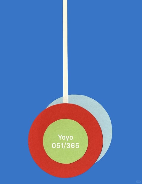 The image presents a minimalist, flat graphic design with a strong illustrative aesthetic, rendered in a clean, vector-like style. The color language is defined by a bold, saturated palette featuring a dominant, vibrant medium blue background, contrasted by a large, circular form composed of concentric rings in a deep, warm red and a muted, light olive green, with a pale, cool blue-grey circle partially visible behind it. A thin, vertical, cream-colored line extends from the top edge to the center of the circular form, suggesting a hanging ornament or pendulum. The lighting is completely flat and uniform, with no discernible shadows or highlights, contributing to a graphic, two-dimensional feel. The texture is smooth and matte, resembling high-quality paper or a digital vector graphic, with no visible grain or surface imperfections. Compositionally, the image is centered and symmetrical along the vertical axis, with the circular element occupying the lower half, creating a balanced and stable arrangement with minimal depth. The shape language is purely geometric, dominated by perfect circles and a straight line, with crisp, clean edges and no organic forms. The overall mood is calm, orderly, and modern, with a playful yet precise energy conveyed through the simple, bold shapes and colors.