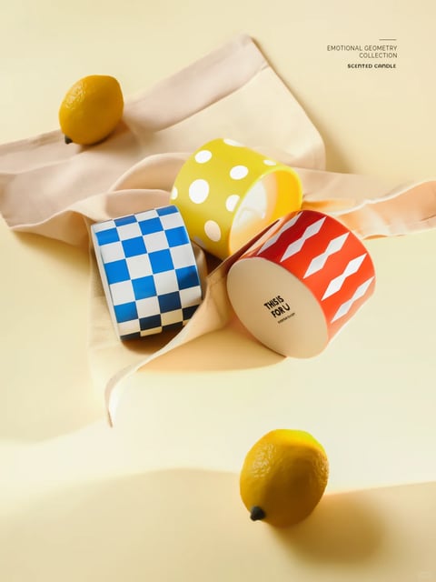 The image presents a photorealistic, product-focused still life with a clean, contemporary aesthetic. The dominant color language is warm and vibrant, featuring a creamy beige background that enhances the high-saturation primary colors of the objects: a cobalt blue and white checkerboard square, a bright lemon-yellow cylinder with white polka dots, and a red cylinder with white diamond patterns. Two real lemons add natural, textured yellow accents. The lighting is soft and diffused, likely from a large, indirect source, creating gentle, elongated shadows that provide depth without harsh contrast, conveying a calm, inviting mood. Textures are rendered with tactile precision: the matte, slightly porous ceramic of the candle holders contrasts with the smooth, waxy skin of the lemons and the soft, flowing drape of the pale pink silk ribbon. Compositionally, the objects are arranged in a dynamic, diagonal cluster on the ribbon, creating a sense of casual elegance and guided visual flow from the top-left lemon to the bottom-right one. The shape language is a deliberate mix of strict geometric forms (squares, circles, diamonds) and the organic, rounded shapes of the lemons and ribbon folds. The overall atmosphere is cheerful, fresh, and minimally sophisticated, evoking a sense of curated, sensory delight.