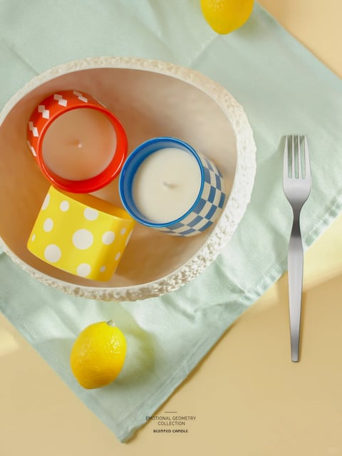 This image presents a high-angle, photorealistic studio photograph with a clean, commercial aesthetic. The color language is dominated by a vibrant, saturated primary palette of red, blue, and yellow, set against a soft, pastel mint-green linen cloth and a warm, pale yellow background, creating a cheerful, high-contrast, and warm-toned composition. The lighting is bright, diffuse, and directional from the upper left, casting soft, defined shadows that enhance the three-dimensionality of the objects without harshness. The texture and material rendering are crisp and tactile, showcasing the glossy, smooth ceramic of the candle containers, the matte, creamy wax, the fine weave of the linen, and the polished, reflective surface of the metal fork. The composition is a carefully arranged flat lay within a circular, textured white bowl, using overlapping shapes and a diagonal placement of the fork to create dynamic balance and depth. The shape language is a playful contrast of strong geometric forms—the cylindrical candles, circular patterns of polka dots and checkerboards—against the organic, irregular edges of the bowl and the natural curves of the lemons. The overall mood is bright, fresh, and whimsically modern, evoking a sense of clean, styled domesticity with a pop-art influence.