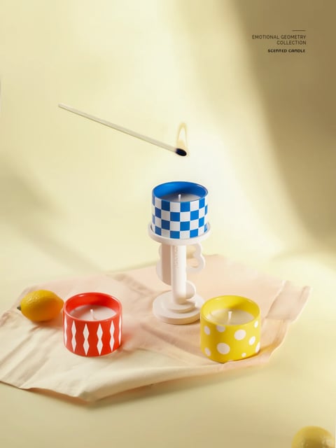 The image presents a photorealistic still life composition with a soft, warm, and inviting aesthetic, rendered with a gentle, diffused quality. The dominant color palette is a vibrant yet harmonious triad of primary colors—bold red, sunny yellow, and deep cobalt blue—set against a creamy, pale yellow background and a soft, off-white linen cloth, creating a cheerful and clean contrast with high saturation and a warm color temperature. Lighting is soft and directional, originating from the upper left, casting long, gentle shadows that add depth and a serene, sunlit mood without harsh edges. The materials are rendered with a smooth, matte finish on the ceramic candle holders and a soft, woven texture on the fabric, emphasizing a tactile, approachable quality. The composition is carefully balanced, with a central elevated blue checkered candle on a white sculptural holder flanked by a red diamond-patterned and a yellow polka-dotted candle, arranged on a casually draped cloth with a lemon adding an organic element, creating a shallow depth of field that focuses attention on the central objects. The shape language is a playful mix of strong geometric patterns (checks, diamonds, dots) on cylindrical forms, contrasted with the organic curves of the lemon and the soft folds of the fabric, all defined by clean, crisp edges. The overall mood is lighthearted, modern, and whimsical, evoking a sense of curated, joyful simplicity with a low-energy, calm visual weight.