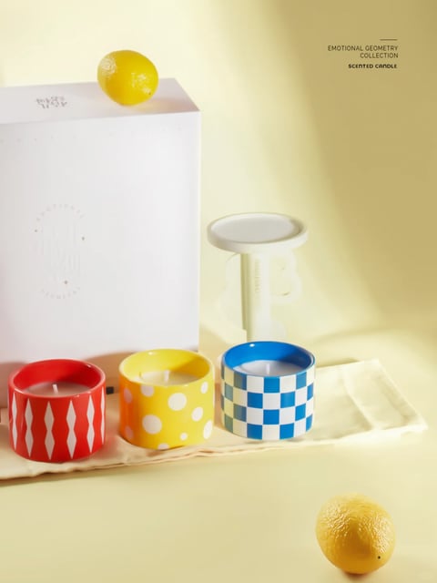 The image presents a photorealistic, studio-lit product composition with a clean, modern aesthetic. The color language is defined by a warm, pale yellow background and surface, contrasted by vibrant, saturated primary colors (red, yellow, blue) in the candle containers and a crisp white box, creating a cheerful, high-contrast palette with a warm overall temperature. Lighting is soft and directional, coming from the upper left, casting gentle, diffused shadows that create depth and a calm, inviting mood. The texture and material feel are smooth and glossy, particularly on the ceramic candle holders and the glossy surface of the lemons, with a subtle matte finish on the white box and a soft, fabric texture on the cloth beneath the candles. The composition is carefully arranged with a shallow depth of field, focusing on the three patterned candles in the foreground, while the white box and a minimalist white candle snuffer recede slightly into the background, framed in a medium shot. Shape language is predominantly geometric, featuring clean circles (candles, lemon), rectangles (box), and bold patterns (diamonds, polka dots, checkerboard), with sharp, defined edges. The overall mood and atmosphere are bright, fresh, and emotionally uplifting, conveying a sense of modern, playful sophistication with a light, airy visual weight.