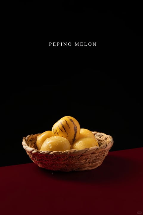The image presents a photorealistic still-life composition featuring a woven wicker basket filled with pepino melons, set against a stark black background and a deep crimson red surface. The visual aesthetic is characterized by a high-contrast, dramatic lighting approach, with a single, directional light source casting sharp highlights and deep, soft-edged shadows, creating a moody and focused atmosphere. The color palette is dominated by the warm, saturated yellows and golden hues of the melons, which contrast vividly with the cool, dark background and the rich, matte red foreground. Textures are rendered with meticulous detail: the basket shows a rough, fibrous weave, while the melons exhibit a smooth, slightly glossy skin with visible water droplets, enhancing their tactile, fresh appearance. Compositionally, the basket is centered in the lower third of the frame, creating a stable, grounded arrangement with a shallow depth of field that keeps the melons sharply in focus. Shape language is primarily organic, with the rounded forms of the melons and the curved lines of the basket contrasting with the geometric, horizontal plane of the surface. The overall mood is elegant, intimate, and slightly theatrical, emphasizing the natural beauty and texture of the subject through controlled lighting and a minimalist setting.