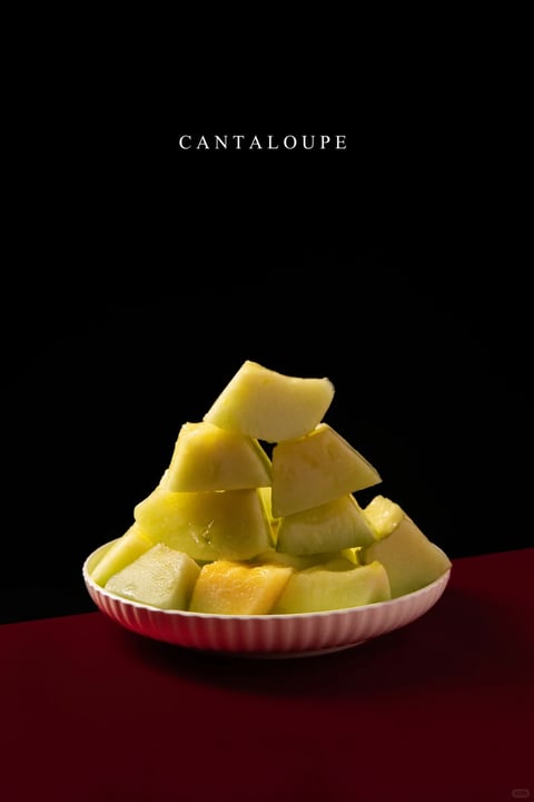 The image presents a photorealistic still life composition of cantaloupe chunks piled in a shallow, fluted pink bowl. The aesthetic is minimalist and high-contrast, with a stark black background that isolates the subject. The color language is defined by the vibrant, warm yellow-green and pale orange flesh of the melon, set against the deep crimson surface below and the void-like black backdrop, creating a bold, limited palette with high saturation on the fruit and deep, muted tones elsewhere. Lighting is directional and hard, coming from the upper left, casting sharp, defined shadows within the pile of fruit and a distinct shadow of the bowl onto the red surface, emphasizing the three-dimensional form and creating a dramatic, focused mood. The texture rendering is highly detailed and tactile, capturing the wet, glistening, fibrous surface of the melon flesh and the smooth, matte finish of the ceramic bowl. Compositionally, the subject is centered in the lower third of a vertical frame, with a pyramidal arrangement of fruit creating a stable, grounded form, and ample negative space above. The shape language is a contrast between the organic, irregular, angular chunks of melon and the perfect, repeating fluted geometry of the bowl's rim. The overall mood is clean, dramatic, and appetizing, with a sense of quiet focus and visual weight concentrated on the textured, illuminated fruit.