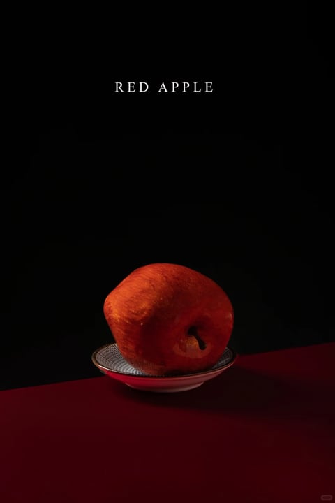 This image features a photorealistic, painterly rendering of a single red apple resting in a shallow, patterned ceramic bowl. The composition is minimalist and dramatically framed against a solid, deep black background, with the apple and bowl placed on a rich, dark red surface that occupies the lower third of the frame. The color language is dominated by a high-contrast, warm palette: the apple exhibits a deep, saturated crimson with subtle orange and yellow undertones where light catches its textured skin, while the bowl shows a delicate blue-and-white geometric pattern with a thin gold rim. Lighting is directional and soft, coming from the upper left, creating gentle highlights on the apple's curve and casting a soft, diffused shadow to the right, enhancing the three-dimensional form and conveying a quiet, contemplative mood. The texture is highly detailed, emphasizing the apple's organic, slightly dimpled skin and the smooth, glazed surface of the ceramic bowl. The shape language is primarily organic, centered on the apple's rounded form, contrasted by the precise, circular geometry of the bowl. The overall atmosphere is one of focused simplicity and dramatic stillness, with the stark black void emphasizing the tactile presence and rich color of the subject.