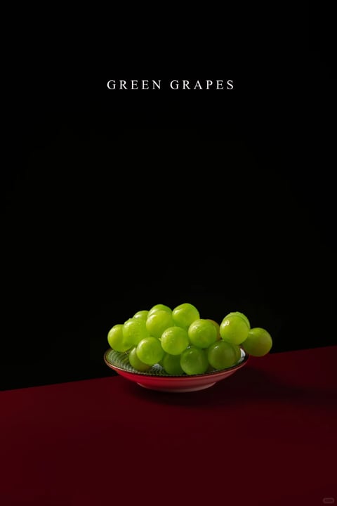 This image features a photorealistic still life composition of green grapes in a shallow bowl, set against a stark black background and resting on a deep maroon surface. The visual aesthetic is clean and minimalist, with a focus on high-contrast lighting and rich color saturation. The color language is dominated by the vibrant, cool green of the grapes, which are rendered with a slight translucency and subtle highlights, contrasting sharply with the deep, warm maroon of the table surface and the absolute black of the background. The lighting approach is dramatic and directional, appearing to come from the upper left, casting soft but defined shadows and creating bright, specular highlights on the grapes' smooth surfaces, emphasizing their roundness and volume. The texture and material feel are highly realistic, with the grapes appearing juicy and firm, their skins smooth with a slight waxy sheen, while the bowl has a glossy, patterned ceramic finish. The composition is centered and balanced, with the bowl of grapes positioned in the lower third of the frame, creating a strong focal point against the negative space above. The shape language is primarily organic, defined by the rounded, clustered forms of the grapes, which contrast with the geometric curve of the bowl's rim. The overall mood and atmosphere are elegant, serene, and slightly dramatic, with a sense of quiet sophistication and visual weight created by the high contrast and deep, rich colors.