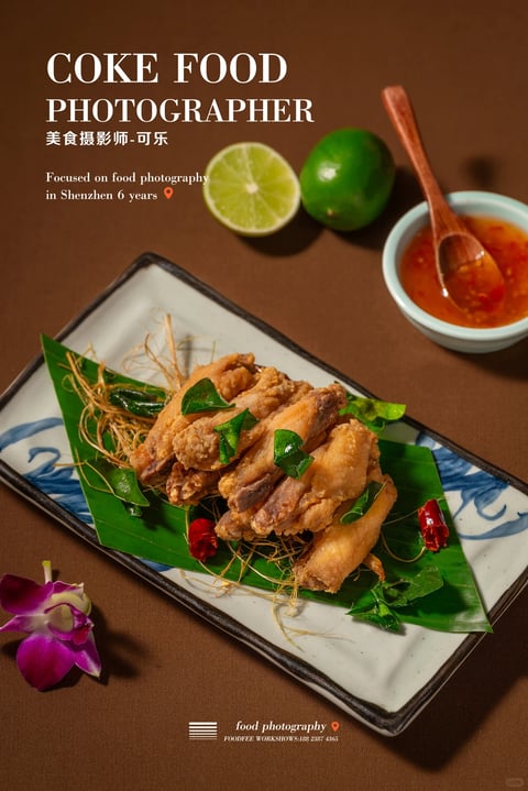 The image presents a photorealistic, high-contrast food photograph with a warm, rich color palette dominated by deep browns, golden ambers, and vibrant greens. The lighting is directional and dramatic, casting soft yet defined shadows that emphasize the crispy, textured surface of the fried chicken wings, which are arranged organically on a green leaf atop a rectangular ceramic plate with blue brushstroke patterns. The composition uses a shallow depth of field, focusing sharply on the foreground food while softly blurring the background elements like the lime halves and dipping sauce, creating a layered spatial arrangement. The texture rendering is highly detailed, highlighting the glistening oil on the chicken, the fibrous quality of the fried garnish, and the smooth, reflective surfaces of the ceramic and liquid sauce. The overall mood is appetizing and sophisticated, with a tactile, inviting atmosphere conveyed through the interplay of warm highlights and cool, deep shadows.