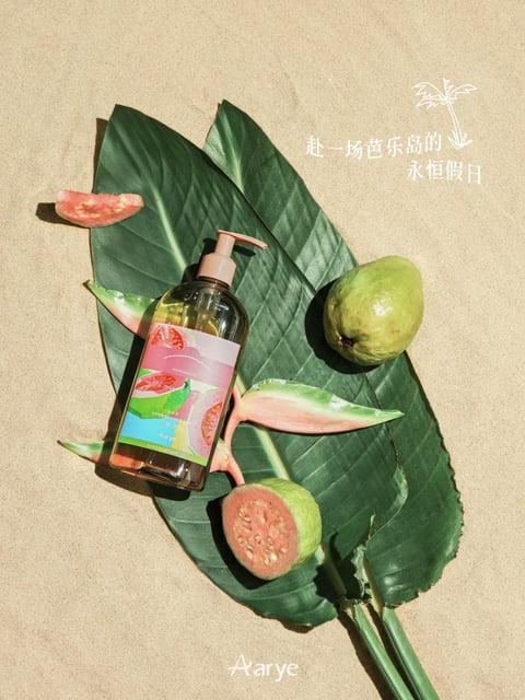 The image presents a product composition on a sandy surface, featuring a clear, cylindrical pump bottle with a peach-colored pump head and a colorful label depicting sliced pink guava. The bottle rests atop two large, dark green, glossy tropical leaves with prominent veins, accompanied by a whole green guava, a halved pink guava, and a small guava slice. The scene is accented by pink and green banana flower bracts, creating a tropical aesthetic. The lighting casts sharp, directional shadows, suggesting bright, direct sunlight. The overall composition is balanced and stylized, with a cohesive color palette of greens, pinks, and sandy beige, evoking a fresh, fruity, and vacation-like atmosphere.