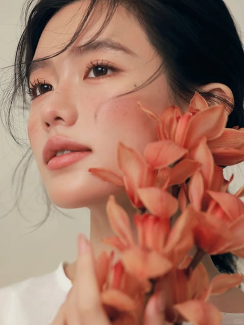 The image presents a photorealistic, high-resolution close-up portrait with a soft, painterly quality in its rendering. The dominant color language is a warm, muted palette centered on peachy-coral and soft terracotta tones, seen in the flowers, blush, and lip color, set against a neutral, off-white background and the subject's dark hair. Saturation is moderate and controlled, with low contrast that creates a gentle, diffused look. The lighting is soft, directional, and appears to come from the upper left, casting subtle, diffused shadows and creating a luminous, glowing effect on the skin with soft highlights on the cheekbone, nose bridge, and lip. Texture and material feel are rendered with a focus on natural, tactile qualities: the skin has a dewy, slightly freckled texture with visible pores and fine hairs, the flower petals appear soft and velvety with delicate veining, and the hair has a fine, wispy texture with individual strands catching the light. The composition is a tight, intimate close-up, framing the subject's face from the forehead to the chin, with the flowers positioned in the foreground, partially obscuring the right side of the face and creating a shallow depth of field where the flowers are slightly softer in focus than the subject's eyes. Shape language is predominantly organic, defined by the soft curves of the facial features, the flowing lines of the hair, and the natural, irregular forms of the flower petals. The overall mood and atmosphere are serene, intimate, and slightly melancholic, with a quiet, contemplative energy and a visual weight that feels delicate and ethereal.