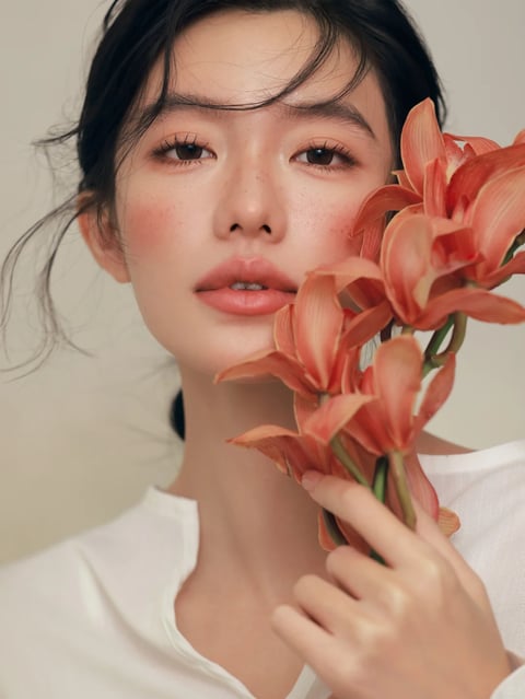 This is a photorealistic close-up portrait with a soft, ethereal quality. The visual aesthetic is defined by a shallow depth of field, rendering the subject's face in sharp detail while the background and parts of the foreground flowers gently blur. The color language is dominated by a warm, muted palette: soft peachy-orange tones in the subject's blush and lip color, complemented by the deeper coral and peach hues of the amaryllis flowers. The skin tones are natural with subtle freckles, and the overall saturation is moderate, leaning towards a soft, desaturated warmth. The lighting is soft and diffused, appearing to come from a large, frontal source, creating gentle, even illumination on the face with minimal harsh shadows, conveying a serene and intimate mood. Texture and material feel are rendered with high fidelity: the delicate, slightly dewy texture of the skin, the fine, wispy strands of dark hair, the soft, matte fabric of the white shirt, and the smooth, slightly waxy surface of the flower petals are all palpable. The composition is a tight, intimate close-up, framing the subject's face from the shoulders up, with the flowers held close to the cheek, creating a sense of proximity and connection. The shape language is predominantly organic, defined by the soft curves of the facial features, the flowing lines of the hair, and the natural forms of the flowers, with soft, blended edges. The mood and atmosphere are calm, tender, and introspective, with a low energy level and a strong visual weight concentrated on the subject's direct gaze and the interplay between the face and the floral element.