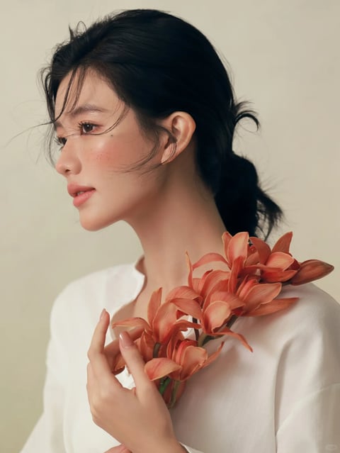 The image presents a photorealistic, studio-style portrait with a soft, painterly rendering technique that emphasizes smooth skin textures and delicate details. The color language is dominated by a warm, muted palette featuring creamy off-white backgrounds, soft peachy-pink blush and lip tones, and deep black hair, with the vibrant coral-orange flowers providing a saturated focal point against the desaturated surroundings. Lighting is soft and diffused, coming from the front-left, creating gentle highlights on the cheekbone and nose with minimal harsh shadows, conveying a serene and intimate mood. Texture and material feel are rendered with high fidelity, showcasing the fine, wispy strands of hair, the subtle matte finish of the skin with faint freckles, the smooth fabric of the white shirt, and the waxy, layered petals of the flowers. Composition is a close-up, off-center profile shot with shallow depth of field, focusing sharply on the face and flowers while the background remains softly blurred. Shape language is predominantly organic, featuring the curved lines of the facial profile, the flowing hair, and the natural forms of the flowers, with soft edge treatment and no hard geometric lines. The overall mood and atmosphere are calm, elegant, and slightly melancholic, with a low-energy, contemplative visual weight.
