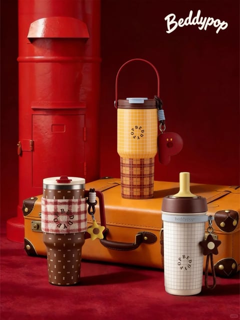 The image displays three insulated tumblers with distinct designs, arranged in a staged, vintage-inspired setting. The tumblers feature a tapered cylindrical form, wider at the top and narrowing towards the base, with a compact, portable silhouette. The left tumbler has a dark brown, glossy finish adorned with a repeating pattern of small, light-colored bone shapes, wrapped in a plush, red-and-white plaid fabric sleeve; its lid is a contrasting cream color with a dark red, flip-up drinking spout, and a yellow star-shaped charm hangs from its side. The central tumbler, positioned on a tan leather suitcase, features a two-tone design: a yellow upper section with a fine white grid pattern and a lower section with a red-and-brown plaid pattern, topped with a dark brown lid and a red carrying handle; a red, heart-shaped charm is attached. The right tumbler has a white, glossy body with a light gray grid pattern, a dark brown lid with a yellow, integrated straw, and a brown star-shaped charm. All tumblers display the circular 'BEDDYPOP' logo. The background includes a tall, glossy red cylindrical structure resembling a vintage postbox, set against a deep red wall and floor, enhancing the warm, nostalgic color palette of reds, browns, yellows, and creams. The lighting creates soft highlights on the glossy surfaces and subtle shadows, emphasizing the textures of the fabric, leather, and metal elements.