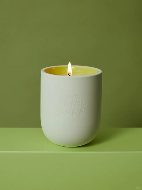 This image presents a photorealistic, studio-style product shot of a single lit candle. The visual aesthetic is clean, minimalist, and modern, with a focus on tactile materiality and serene ambiance. The color language is dominated by a cohesive, monochromatic green palette, ranging from a soft, muted sage green in the background to a slightly more saturated, warm olive green in the foreground surface, creating a subtle tonal shift. The candle wax itself is a vibrant, translucent chartreuse yellow, providing a focal point of saturated color. The lighting is soft and diffused, appearing to come from the upper left, casting gentle, elongated shadows to the right and below the candle, which enhances its three-dimensional form without creating harsh contrasts. The texture and material feel are distinctly rendered: the candle holder is a matte, unglazed ceramic with a smooth, slightly porous surface, while the wax appears glossy and semi-translucent where melted near the flame. The composition is centered and balanced, with the candle placed on a flat plane against a seamless backdrop, creating a shallow depth of field that keeps the subject sharply in focus. The shape language combines the soft, organic curves of the rounded candle holder and the flickering flame with the clean, geometric lines of the embossed text ('LOVE FLORAL DRAMA') on the holder's surface. The overall mood and atmosphere are calm, tranquil, and slightly intimate, evoking a sense of quiet warmth and focused simplicity.