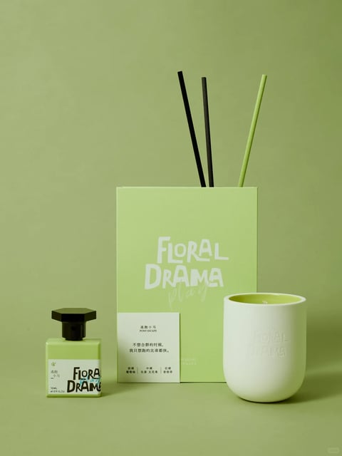The image presents a product photography composition with a cohesive, monochromatic green aesthetic, rendered in a clean, photorealistic style. The dominant color palette is a muted, desaturated sage green, used for the background, product packaging, and diffuser reeds, creating a soft, low-contrast, and cool-toned visual field. Lighting is soft and diffused, appearing to come from the front-left, casting gentle, subtle shadows that define form without harshness, conveying a calm and serene mood. Textures are smooth and matte, with the paper packaging showing a fine grain, the ceramic candle holder a soft, non-reflective finish, and the glass perfume bottle a subtle sheen, all emphasizing a tactile, premium feel. The composition is balanced and centered, with the tall rectangular box as the vertical anchor, flanked by the shorter, squatter perfume bottle on the left and the rounded candle holder on the right, creating a stable, symmetrical arrangement with clear spatial separation. Shape language is predominantly geometric, featuring clean rectangles and cylinders with sharp edges, contrasted by the organic, hand-drawn typography on the packaging. The overall mood is minimalist, tranquil, and sophisticated, with a low-energy, contemplative atmosphere emphasized by the unified color scheme and soft lighting.