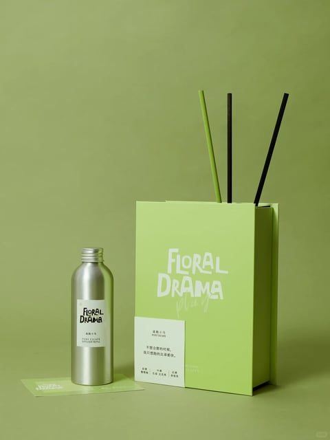 This image presents a photorealistic, studio-shot still life with a clean, minimalist aesthetic. The dominant color palette is a cohesive range of muted, desaturated greens, from the soft sage-green background to the brighter, almost lime-green paper bag, creating a monochromatic and calming atmosphere. The lighting is soft and diffused, likely from a large, frontal source, casting gentle, subtle shadows that define form without harsh contrast, contributing to a serene and modern mood. The materials are rendered with high fidelity: the matte, slightly textured surface of the paper bag contrasts with the smooth, brushed metallic finish of the aluminum bottle and the fibrous, porous texture of the diffuser sticks. Compositionally, the arrangement is asymmetrical yet balanced, with the vertical bag and sticks offset by the shorter bottle, creating a sense of organized simplicity. The shape language is predominantly geometric and clean, featuring sharp rectangular forms, cylindrical volumes, and straight lines, with crisp edges and minimal ornamentation. The overall atmosphere is one of quiet sophistication, tranquility, and contemporary design.