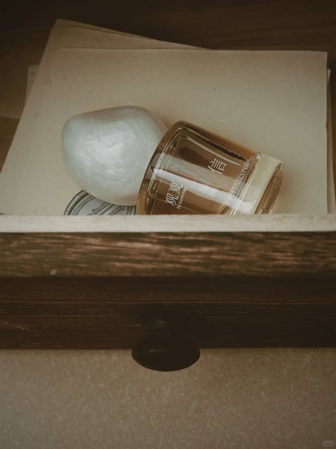 The image presents a photorealistic, close-up still life with a muted, desaturated color palette dominated by warm beiges, soft creams, and earthy browns, punctuated by the amber-gold liquid in a glass bottle and the pearlescent white of a smooth, organic-shaped stone. The lighting is soft and diffused, appearing to come from the upper left, casting gentle, subtle shadows that create a quiet, intimate mood. Textures are rendered with tactile detail: the grainy, fibrous surface of aged paper, the smooth, cool glass with its etched white text, the polished, iridescent stone, and the rough, weathered wood grain of the drawer front. The composition is tightly framed within the drawer, arranging the objects in a casual, overlapping cluster that suggests a personal, stored-away collection, with a shallow depth of field focusing on the central items. Shapes contrast between the geometric, cylindrical bottle and the organic, rounded forms of the stone and drawer handle, with clean edges on the manufactured items against the softer contours of natural materials. The overall atmosphere is nostalgic, calm, and tactile, evoking a sense of quiet contemplation and preserved memory.