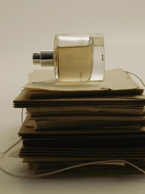 This image presents a photorealistic still life with a minimalist, serene aesthetic. The composition centers on a clear, rectangular glass perfume bottle with a silver spray nozzle, lying horizontally atop a stack of aged, textured paper envelopes. The color language is muted and warm, dominated by creamy off-whites, soft beiges, and the pale amber hue of the liquid inside the bottle, with subtle metallic silver accents. The lighting is soft and diffused, appearing to come from the upper left, casting gentle, elongated shadows that enhance the three-dimensionality of the objects without harsh contrast. Textures are rendered with high fidelity: the smooth, reflective surface of the glass contrasts with the rough, fibrous, and slightly uneven edges of the stacked paper, which shows visible layers and a tactile, handmade quality. The composition is tightly framed, focusing on the horizontal arrangement of the bottle and the vertical stack of paper, creating a balanced, stable structure with a shallow depth of field that keeps the subjects sharp against a plain, neutral background. Shapes are primarily geometric and clean—the rectangular bottle and stacked papers—juxtaposed with the organic, frayed edges of the paper and the delicate, curved string trailing from the stack. The overall mood is calm, quiet, and contemplative, evoking a sense of nostalgia and understated elegance.
