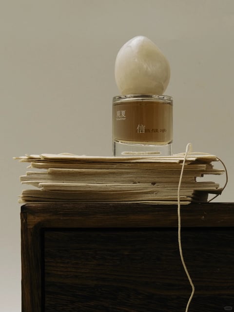 This image is a photorealistic still life with a soft, painterly quality, rendered with high detail and a tactile sense of materiality. The dominant color palette is muted and warm, featuring creamy off-whites, aged beige, and rich amber-brown tones, with low saturation and gentle contrast that creates a cohesive, earthy feel. Lighting is soft and directional, appearing to come from the upper left, casting subtle, diffused shadows that enhance the three-dimensionality of the objects without harshness, conveying a calm, contemplative mood. Textures are rendered with exceptional detail: the stacked papers show frayed, deckled edges and a fibrous, matte surface; the glass bottle is smooth and transparent with subtle reflections; the stone-like cap has a polished, slightly irregular organic texture; and the dark wood surface below has a visible grain and a matte finish. The composition is a centered, eye-level shot with a shallow depth of field, focusing sharply on the bottle and the top of the paper stack while the background is a plain, out-of-focus neutral wall. Shape language combines organic forms (the egg-shaped cap, the uneven paper edges) with geometric precision (the cylindrical glass bottle, the rectangular wood block), all defined by clean, soft edges. The overall mood is serene, nostalgic, and grounded, with a quiet, meditative atmosphere emphasized by the harmonious arrangement and subdued, natural tones.