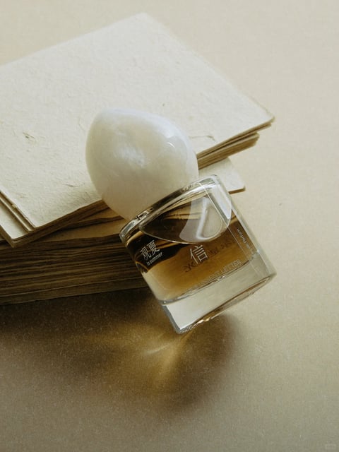This is a photorealistic, still-life photograph with a minimalist, elegant aesthetic. The dominant color language is a warm, monochromatic palette of creamy beiges, soft tans, and amber-gold tones, with low saturation and gentle contrast. The lighting is soft and diffused, appearing to come from the upper left, casting subtle, elongated shadows that create a calm, serene mood. The image features distinct textures: the rough, fibrous surface of stacked, handmade paper sheets contrasts with the smooth, glossy, translucent glass of a perfume bottle and its opaque, polished, egg-shaped white cap. The composition is a close-up, slightly high-angle shot with a shallow depth of field, focusing sharply on the bottle while the background paper softly blurs. The shape language combines the organic, irregular edges of the paper stack and the curved cap with the clean, geometric lines of the rectangular glass bottle. The overall atmosphere is tranquil, sophisticated, and tactile, evoking a sense of quiet luxury and natural simplicity.