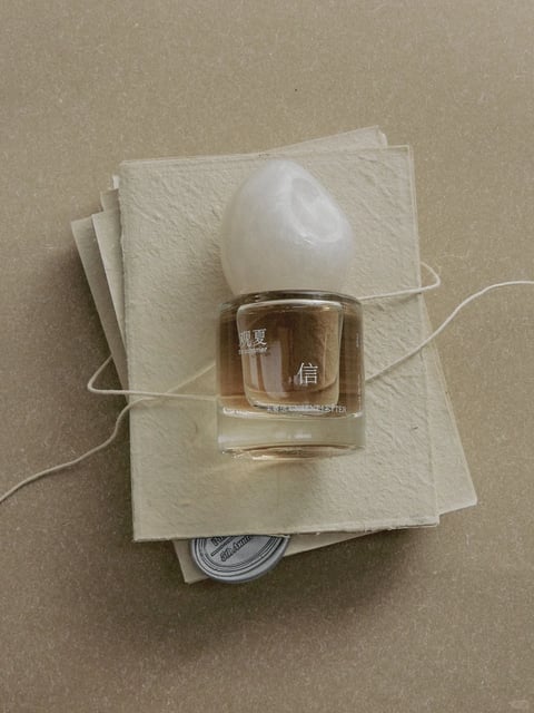 The image presents a photorealistic, top-down still life with a minimalist, tactile aesthetic. The dominant color language is a muted, earthy palette of warm beige, cream, and soft brown tones, with low saturation and gentle contrast that creates a calm, natural temperature. Lighting is soft and diffused from an overhead direction, casting subtle, soft-edged shadows that enhance the three-dimensionality of the objects without harshness. Texture and material feel are central, with the rough, fibrous surface of the handmade paper contrasting against the smooth, glossy glass bottle and its matte, sculptural white cap. Compositionally, the objects are arranged in a centered, layered stack on a flat surface, creating a shallow depth of field that emphasizes the central bottle. Shape language blends organic forms, like the irregular paper edges and the curved, pebble-like bottle cap, with the clean, geometric cylinder of the glass. The overall mood is serene, intimate, and nostalgic, evoking a sense of quiet contemplation and artisanal craftsmanship.