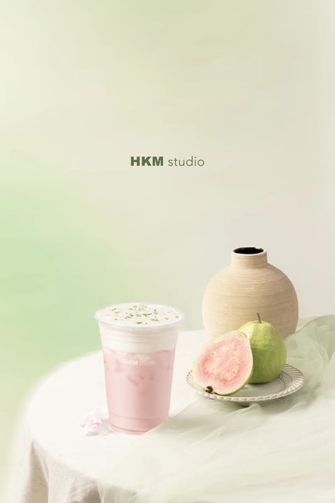The image presents a photorealistic, studio-style composition with a soft, diffused aesthetic. The dominant color palette is a muted, pastel scheme centered on a gradient background that transitions from a pale, creamy white to a soft, desaturated mint green, creating a gentle, cool-toned atmosphere. The lighting is soft and directional, appearing to come from the upper left, casting subtle, diffused shadows that give the objects a gentle volume without harsh contrast. The textures are rendered with a focus on tactile realism: the smooth, matte ceramic of the beige vase, the slightly rough, speckled surface of the cut guava, and the creamy, frothy top layer of the pink beverage. The composition is asymmetrical and balanced, with the beverage cup positioned in the foreground left, the plate of fruit in the mid-ground right, and the vase anchoring the background right, creating a clear sense of depth through overlapping forms. The shape language is predominantly organic, featuring the rounded curves of the cup, fruit, and vase, contrasted only by the clean, geometric lines of the plate's rim. The overall mood is serene, fresh, and minimalist, evoking a calm, clean, and slightly luxurious feeling through its restrained color use and elegant simplicity.
