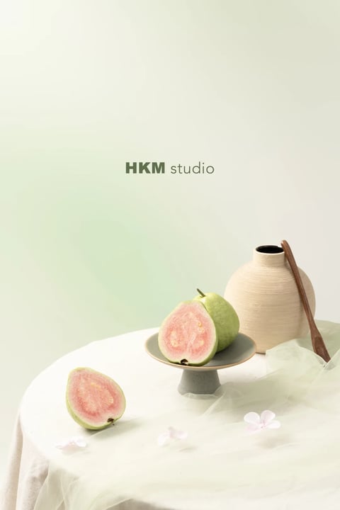 This image presents a minimalist still life rendered in a soft, photorealistic style with a gentle, painterly quality. The dominant color palette is muted and harmonious, featuring a gradient background transitioning from a pale sage green at the top to an off-white at the bottom, complemented by the soft pink flesh and pale green rind of the guavas, the warm beige of the ceramic vase, and the earthy brown of the wooden spoon. The color temperature is neutral to slightly cool, with low saturation and low contrast, creating a serene and airy feel. Lighting is soft and diffused, appearing to come from the upper left, casting gentle, subtle shadows that define forms without harshness, conveying a calm, natural daylight mood. Textures are rendered with a tactile, organic quality: the matte, slightly porous surface of the unglazed ceramic, the smooth yet subtly speckled skin of the guava, the soft drape of the sheer fabric, and the fine grain of the wooden spoon. The composition is asymmetrical and balanced, with the subjects arranged on a round table draped in white linen and a sheer green overlay, creating a shallow depth of field where the foreground guava slice is slightly softer than the central arrangement. Shape language is predominantly organic, with the rounded forms of the fruit, vase, and table contrasted by the gentle, flowing lines of the fabric and the straight handle of the spoon, all rendered with soft, blended edges. The overall mood is tranquil, fresh, and elegantly simple, evoking a sense of quiet abundance and natural beauty with very low visual energy and weight concentrated in the lower right quadrant.