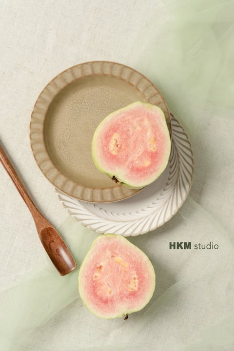 This image presents a photorealistic, minimalist still life with a soft, airy aesthetic. The color language is dominated by a muted, natural palette: the pale, desaturated sage green of the sheer fabric and guava rind contrasts gently with the vibrant yet soft coral-pink of the fruit's flesh, all set against a warm, neutral beige linen background. The lighting is soft and diffused, appearing to come from the upper left, casting gentle, elongated shadows that create a calm, serene mood without harsh contrast. Textures are rendered with high fidelity: the coarse weave of the linen, the delicate, translucent drape of the tulle, the matte, slightly speckled ceramic glaze of the stacked plates, and the moist, granular surface of the cut fruit are all tactile and detailed. The composition is a balanced, overhead flat lay, with the stacked plates and guava halves creating a central focal point, while the wooden spoon and fabric folds guide the eye through the frame. Shapes are predominantly organic, seen in the rounded fruit and curved spoon, contrasted by the geometric, fluted edges of the plates. The overall mood is tranquil, fresh, and elegantly rustic, conveying a sense of quiet, natural beauty.