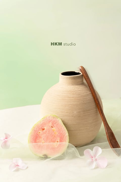 The image presents a photorealistic still life with a soft, painterly aesthetic, characterized by a gentle, diffused lighting approach that casts subtle, soft-edged shadows, creating a serene and airy atmosphere. The dominant color palette is a muted, desaturated blend of pale mint green, warm beige, and soft pink, with a low-contrast, high-key tonal range that emphasizes a calm, natural mood. Textures are rendered with tactile clarity: the ribbed, matte surface of the ceramic vase, the smooth, fibrous flesh of the pink guava, the delicate, sheer tulle fabric, and the polished grain of the wooden spoon. The composition is asymmetrical and balanced, with a shallow depth of field that keeps the central objects in focus against a softly blurred background, using organic shapes and soft edges to create a harmonious, tranquil visual weight.