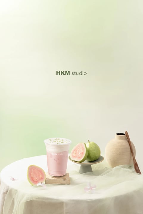 The image presents a photorealistic, studio-lit still life with a soft, painterly quality, characterized by a dominant palette of muted, desaturated pastels—primarily pale sage green, blush pink, and creamy off-white—with low saturation and gentle contrast. The lighting is soft and diffused, originating from the upper left, casting subtle, elongated shadows that create a calm, serene mood. Textures are rendered with tactile detail: the matte, slightly porous surface of a ceramic vase, the smooth, translucent quality of a sheer fabric drape, the creamy, frothy top of a pink beverage, and the grainy, seeded flesh of sliced pink guavas. The composition is asymmetrical yet balanced, with objects arranged on a circular table draped in fabric, creating a shallow depth of field that emphasizes the central drink and fruit while softly blurring the background. Shapes are predominantly organic, with the rounded forms of the guavas, vase, and cup contrasting against the subtle geometric lines of the table edge and a small rectangular stone coaster. The overall mood is tranquil, fresh, and aesthetically refined, evoking a sense of gentle, natural elegance.