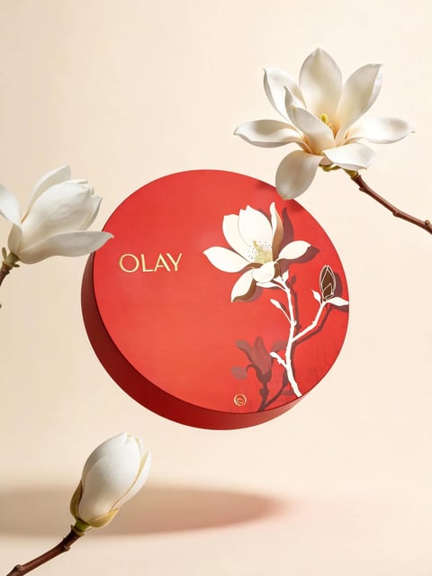 The image presents a centered, circular red product box against a soft, uniform beige background, creating a minimalist and elegant composition. The box, the main subject, is flanked by three realistic white magnolia flowers on slender brown branches: one fully bloomed flower in the upper right, another partially opened bloom on the left, and a closed bud in the lower left, adding organic framing and depth. The visual aesthetic is a blend of photorealistic floral elements with a stylized, graphic illustration on the box itself. The color language is dominated by a vibrant, saturated crimson red on the box, contrasted by the pure white of the magnolias and the warm, neutral beige backdrop, with gold accents providing a touch of luxury. Lighting is soft and diffused, appearing to come from the upper left, casting gentle, realistic shadows that give the objects volume and a subtle three-dimensionality. The texture and material feel are distinct: the real flowers have a delicate, waxy petal texture and woody branches, while the box surface appears smooth and matte with a printed, slightly stylized floral graphic. Compositionally, the circular box is the focal point, with the surrounding flowers creating a balanced, asymmetrical arrangement that guides the eye inward. Shape language combines the perfect geometric circle of the box with the organic, flowing lines of the magnolia branches and petals. The mood is serene, sophisticated, and celebratory, evoking a sense of premium beauty and natural elegance. Visible text includes the word 'OLAY' in a clean, sans-serif, gold-colored font positioned on the upper left quadrant of the red box, serving as clear brand identification.