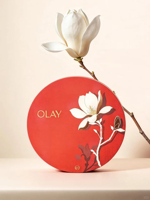 The image features a minimalist, elegant composition centered on a large, circular red object, likely a decorative box or lid, resting on a smooth, pale beige surface against a matching background. A single, real white magnolia flower on a slender brown branch extends diagonally from the upper right, arching over the red circle, creating a visual dialogue between the three-dimensional natural element and the two-dimensional illustrated design on the circle's surface. The red circle is adorned with a stylized, painterly illustration of a white magnolia bloom and branch in white and brown tones, positioned on the right side, with subtle, darker shadow-like forms beneath it. The visual aesthetic blends photorealistic rendering of the real flower and branch with a flat, illustrative style for the graphic on the red surface, creating a sophisticated, mixed-media feel. The color language is dominated by a warm, saturated, festive red for the central object, contrasted sharply by the pure white of the magnolia petals and the soft, neutral beige of the environment, with accents of brown in the branches and gold in the text. The lighting is soft, diffused, and directional from the upper left, casting gentle shadows that give the real flower volume and ground the red circle, contributing to a serene, calm mood. Texture and material are clearly differentiated: the red circle has a matte, slightly textured paper or cardboard finish, the real flower petals appear velvety and delicate, the branch looks woody and rough, and the surface beneath is smooth and seamless. The composition is balanced yet asymmetrical, with the heavy visual weight of the red circle anchored in the lower center, counterbalanced by the lighter, reaching branch above, using negative space effectively. Shape language contrasts the perfect geometric circle with the organic, flowing curves of the magnolia petals and branches. The overall mood is one of tranquil elegance, purity, and refined beauty, with a festive yet understated atmosphere. The only visible text is the word 'OLAY' printed in a clean, gold, sans-serif font on the left side of the red circle, serving as a brand identifier integrated into the design.
