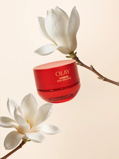 The image presents a photorealistic, high-end product shot featuring a vibrant red Olay Peptides cream jar as the central subject, suspended elegantly between two pristine white magnolia blossoms on a slender brown branch. The composition is balanced and airy, with the jar positioned in the mid-ground, framed by one magnolia bloom above and another below, against a soft, uniform cream-beige background that creates a serene, minimalist atmosphere. The visual aesthetic is clean and luxurious, with a focus on realistic textures and soft, diffused lighting. The color language is dominated by the saturated, glossy red of the jar, contrasted with the pure white petals and the warm neutral backdrop, creating a striking yet harmonious palette with high contrast between the red and white. The lighting is soft and directional, appearing to come from the upper left, casting gentle highlights on the jar's curved surface and the petals' edges, enhancing their three-dimensional form without harsh shadows. Texture and material feel are rendered with precision: the jar exhibits a smooth, reflective glass-like finish with a subtle gradient, while the magnolia petals appear velvety and delicate, and the branch shows a natural, slightly rough bark texture. Compositionally, the arrangement uses negative space effectively, with the diagonal line of the branch guiding the eye through the frame, and the floating jar creating a sense of weightlessness and focus. Shape language combines the organic, flowing curves of the flowers and branch with the clean, geometric silhouette of the cylindrical jar, its edges softly rounded. Visible text on the jar includes the brand name 'OLAY' in elegant gold serif font, with 'PEPTIDES' in smaller uppercase letters below, and Chinese characters '紧致肽·淡纹肽·信号肽' (translating to 'Firming Peptide · Anti-wrinkle Peptide · Signal Peptide') beneath that, all centered on the jar's lid, reinforcing the product's identity and key ingredients. The overall mood is one of refined elegance, tranquility, and premium skincare, with a calm, uplifting energy conveyed through the soft lighting and natural elements.