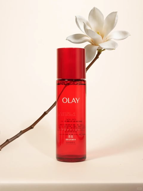 The image presents a photorealistic product photograph of a single Olay skincare bottle, centrally positioned against a soft, neutral beige background. The composition is clean and minimalist, with the bottle as the clear focal point, accompanied by a single, elegant branch of a white magnolia flower that arches gracefully from the lower left, passing behind the bottle and extending towards the upper right. The bottle itself is a tall, cylindrical container made of translucent red glass or plastic, filled with a clear liquid, and topped with a solid, glossy red cap. The overall aesthetic is one of refined elegance and natural purity. The color language is dominated by the vibrant, saturated crimson of the bottle, which creates a strong contrast against the muted, warm beige of the background and the pristine white of the magnolia petals. The lighting is soft and diffused, appearing to come from the front-left, casting gentle, subtle shadows to the right and behind the objects, which enhances the three-dimensional form without creating harsh lines. The texture and material rendering are highly detailed: the bottle exhibits a smooth, glossy surface with subtle reflections, the liquid inside shows tiny bubbles at the meniscus, the flower petals have a delicate, matte, velvety appearance, and the branch has a rough, woody texture with visible nodes. The composition follows a balanced, vertical arrangement with a strong diagonal element introduced by the flower branch, creating dynamic visual interest while maintaining a sense of calm. The shape language combines the clean, geometric cylinder of the bottle with the organic, flowing curves of the flower petals and the irregular line of the branch. The mood and atmosphere conveyed are serene, sophisticated, and fresh, emphasizing the product's connection to natural beauty and effective skincare. Visible text is printed in white on the front of the bottle. The most prominent text is the brand name 'OLAY' in a clean, sans-serif font, positioned in the upper third of the bottle's body. Below this, smaller text in both English and Chinese characters is visible, though partially obscured by the liquid's refraction and the branch; readable elements include 'YAJO' (likely a reversed 'OLAY' seen through the bottle), Chinese characters, and the volume indication '150ml' near the bottom. This text serves a branding and informational role, identifying the product and its specifications.