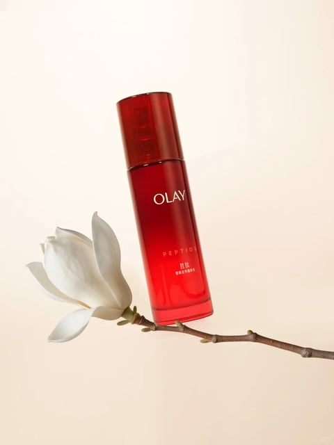 The image features a single, tall, cylindrical red bottle of Olay skincare product as the central subject, positioned slightly off-center and leaning against a slender, dark brown branch with a single, large, white magnolia blossom. The composition is minimalist and clean, set against a smooth, uniform, pale beige or off-white background that creates a soft, neutral canvas. The bottle has a glossy, translucent red finish with a matching red cap, and its surface reflects light subtly, suggesting a smooth, glass-like material. Visible text on the bottle includes the brand name 'OLAY' in white, uppercase sans-serif font near the top, the word 'PEPTIDE' in smaller white uppercase letters below it, and Chinese characters '胜肽' and '塑颜金纯精华乳' in even smaller white text at the bottom, indicating the product line and type. The lighting is soft and diffused, coming from the upper left, casting gentle shadows to the right and below the objects, which enhances the three-dimensional form without harsh contrasts. The color palette is dominated by the vibrant, saturated red of the bottle, contrasted sharply by the pure white of the magnolia flower and the muted brown of the branch, all set against the warm, pale background. The texture contrast is notable: the smooth, hard, reflective surface of the bottle versus the soft, matte, organic petals of the flower and the rough, woody texture of the branch. The overall mood is elegant, serene, and sophisticated, with a focus on clean lines, natural elements, and a sense of calm luxury.