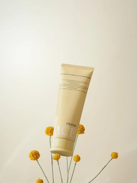 The image features a single, upright, beige-colored tube of skincare product as the central subject, positioned against a plain, off-white background. The tube is supported by several thin, green stems topped with small, round, yellow, pom-pom-like flowers (resembling craspedia or billy buttons), which emerge from the bottom of the frame. The composition is minimalist and centered, with a shallow depth of field that keeps the product and flowers in sharp focus while the background remains soft and neutral. The visual aesthetic is clean, modern, and photorealistic, with a focus on product presentation. The color palette is dominated by warm, muted tones: the soft beige of the tube, the vibrant yet natural yellow of the flowers, and the pale green stems, all set against a creamy off-white backdrop, creating a low-contrast, harmonious, and warm color temperature. The lighting is soft, diffused, and appears to come from the front-left, casting gentle, subtle shadows that define the tube's cylindrical form and the texture of the flower heads without creating harsh lines, conveying a calm and gentle mood. The texture and material feel are rendered with clarity: the tube has a smooth, matte plastic finish with a slight sheen at the curved edges, while the flower heads exhibit a dense, fuzzy, or granular texture. The composition uses a vertical orientation with the product slightly angled, creating a dynamic yet balanced arrangement where the organic, spherical shapes of the flowers contrast with the geometric, cylindrical shape of the tube. The visible text on the tube is in English, printed in a clean, sans-serif font. Near the top, it reads "CLEAR ESSENCE ANTI-WRINKLE 2-IN-1 ICE CREAM CLEANSER" followed by smaller descriptive text about ingredients and benefits. Lower on the tube, the brand name "YOUZISE" and the large, stylized word "ACE" are printed in a lighter, possibly silver or white, font, serving as the primary branding and product identification elements. The overall mood is serene, natural, and focused on gentle skincare, with a tactile quality emphasized by the contrast between the smooth tube and the textured botanical elements.