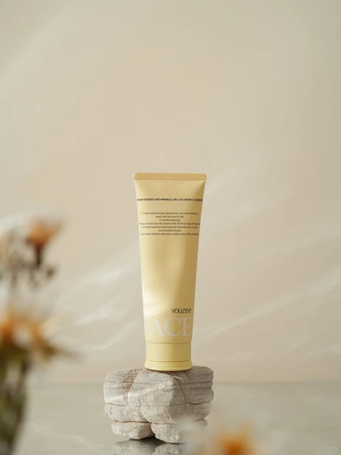 The image presents a photorealistic product shot centered on a single tube of facial cleanser, positioned vertically on a rough, layered stone pedestal against a soft, neutral beige background. The composition is minimalist and balanced, with the tube as the clear focal point, supported by the textured stone base and framed by out-of-focus, warm-toned botanical elements in the foreground that add depth and a natural, organic feel. The tube itself is a soft, matte yellow with a slightly tapered shape and a crimped top edge, featuring black and white text. The readable text includes the product name 'CAVIAR ESSENCE ANTI-WRINKLE 2-IN-1 ICE CREAM CLEANSER' at the top, followed by numbered benefits in English describing its moisturizing, gentle cleaning, and caviar essence properties, and the brand name 'YOUZISE' with the word 'FACE' prominently displayed in large, white, serif font near the bottom. The lighting is soft and diffused, appearing to come from the upper left, casting gentle shadows to the right and creating a warm, serene, and premium atmosphere. The color palette is dominated by warm, muted tones—soft yellows, creamy beiges, and earthy browns—with low contrast and a warm color temperature, evoking a sense of calm and natural luxury. The textures are carefully rendered: the smooth, matte plastic of the tube contrasts with the rough, porous, and layered surface of the stone pedestal, while the blurred botanicals suggest a delicate, dry texture. The overall aesthetic is clean, modern, and aspirational, with a focus on tactile materiality and a gentle, inviting mood.