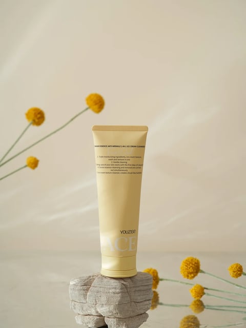 The image is a photorealistic product photograph featuring a single beige tube of facial cleanser as the main subject, positioned vertically on a rough, light gray stone pedestal. The scene is minimalist and serene, with a soft, warm, and neutral color palette dominated by creamy beiges, soft yellows, and muted grays. The background is a smooth, out-of-focus gradient of pale beige and cream, creating a clean, studio-like setting. The composition is centered and balanced, with the product tube as the clear focal point, framed by delicate, blurred yellow craspedia (billy button) flowers on thin green stems in the foreground and background, adding a touch of organic softness and depth. The lighting is soft and diffused, appearing to come from the upper left, casting gentle, subtle shadows that enhance the three-dimensionality of the tube and stone without creating harsh contrast. The visual aesthetic is clean, modern, and elegant, with a focus on tactile textures: the smooth, matte plastic of the tube contrasts with the rough, layered, and porous surface of the stone pedestal. The tube features visible text in a clean, sans-serif font; near the top, it reads 'CAVIAR ESSENCE ANTI-WRINKLE 2-IN-1 ICE CREAM CLEANSER' in small, dark print, followed by a numbered list of product benefits (e.g., '1. Triple moisturizing ingredients...'), and at the bottom, the brand name 'YOUZISE' and the partial word 'FACE' in larger, embossed or subtly printed lettering. The overall mood is calm, sophisticated, and natural, evoking a sense of gentle, effective skincare.