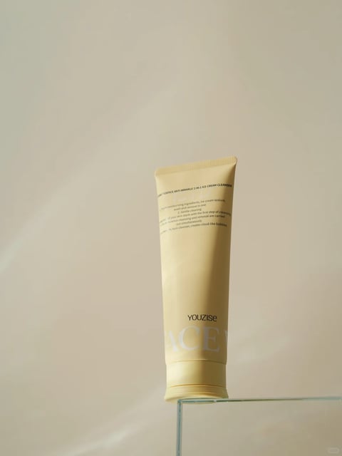 The image presents a single, vertically oriented tube of facial cleanser, positioned slightly off-center on the edge of a clear, minimalist glass surface against a seamless, warm beige background. The composition is clean and focused, utilizing negative space to emphasize the product. The tube itself is a soft, pale yellow with a matte finish, featuring a crimped top seal and a screw-on cap at the base. Visible text on the tube includes the brand name 'YOUZISE' in a clean, black sans-serif font near the bottom, with the partial word 'ACE' in large, faint white letters below it. Further up, smaller black text reads '...ESSENCE ANTI-WRINKLE 2-IN-1 ICE CREAM CLEANSER' and descriptive phrases like 'moisturizing ingredients, ice cream texture,' 'Gentle cleaning,' and 'creates cloud-like bubbles.' The lighting is soft and diffused, coming from the upper left, casting a gentle, elongated shadow to the right and creating a subtle highlight along the left edge of the tube, enhancing its cylindrical form. The overall aesthetic is photorealistic with a minimalist, serene mood, characterized by a warm, monochromatic color palette of beige and pale yellow, low contrast, and a smooth, tactile material feel that suggests a creamy, gentle product.
