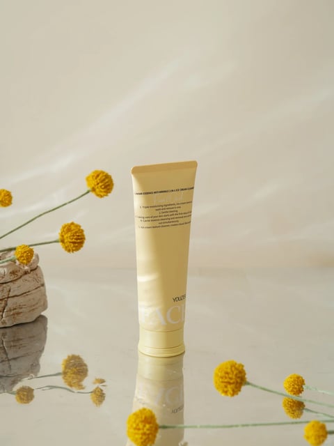 The image presents a minimalist, photorealistic still life composition centered on a single, upright tube of skincare product. The main subject is a pale yellow, matte-finish tube with a crimped top, positioned vertically on a highly reflective, mirror-like surface that creates a clear, slightly distorted reflection of the tube and surrounding elements. To the left, a rough, textured, light brown stone or clay vessel holds several stems of yellow craspedia (billy button) flowers, whose round, pom-pom-like heads add organic, spherical shapes. Additional craspedia stems are scattered on the reflective surface in the foreground and background, creating a sense of depth and framing the central product. The background is a seamless, soft, warm beige or off-white, with a subtle gradient and a faint, diagonal shadow cast across the upper portion, suggesting directional light from the upper left. The overall color palette is monochromatic and warm, dominated by shades of pale yellow, cream, beige, and the golden yellow of the flowers, with low saturation and soft contrast. The lighting is soft and diffused, creating gentle highlights on the tube's curve and the flower heads, and long, soft shadows, evoking a calm, serene, and natural atmosphere. Visible text on the tube is printed in a small, dark, sans-serif font. At the top, it reads "CAVIAR ESSENCE ANTI-WRINKLE 2-IN-1 ICE CREAM CLEANSER". Below this, a numbered list describes product benefits, with phrases like "Triple moisturizing ingredients," "wash and nourish," and "ice cream texture cleanser" partially legible. Near the bottom, the brand name "YOUZISE" is printed above the large, partially obscured word "FACE" in a bold, white, sans-serif font. The text serves a functional, informational role, detailing the product's identity and claims. The focus is sharp on the product tube and the flowers immediately adjacent to it, with a shallow depth of field that gently softens the foreground and background elements, guiding the viewer's eye to the central subject. The texture of the tube is smooth and matte, contrasting with the rough, porous surface of the stone vessel and the dense, fuzzy texture of the flower heads. The composition is balanced and clean, using negative space and reflective symmetry to create a sophisticated, commercial aesthetic focused on natural ingredients and gentle skincare.