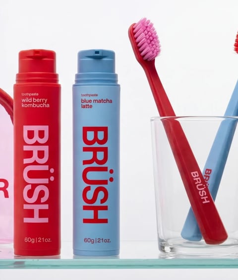This is a bright, high-key product photograph showcasing two cylindrical toothpaste tubes and two matching toothbrushes, arranged on a glass shelf against a clean white background. The composition is balanced and symmetrical, with the red tube on the left and the light blue tube on the right, each paired with a toothbrush of the same color placed in a clear glass tumbler to the right. The overall visual aesthetic is modern, minimalist, and photorealistic, with a focus on clean lines and bold, graphic design. The color language is defined by a vibrant, high-saturation palette of primary red and soft, pastel sky blue, creating a strong, playful contrast. The lighting is soft, diffuse, and even, coming from the front, which minimizes harsh shadows and gives the products a smooth, approachable feel. The texture and material feel are rendered with precision: the toothpaste tubes have a matte, slightly textured plastic finish, while the toothbrush handles appear smooth and glossy, and the glass tumbler is perfectly transparent with subtle reflections. The composition uses a shallow depth of field, keeping the products in sharp focus while the background remains a clean, undistracted white. The shape language is predominantly geometric and cylindrical, with clean, straight lines and rounded edges, emphasizing a modern, functional design. The mood is fresh, clean, and energetic, conveying a sense of contemporary oral care with a fun, trendy twist. The visible text is integral to the design: on the red tube, the words 'toothpaste', 'wild berry kombucha', and the large vertical brand name 'BRÜSH' are printed in white; on the blue tube, the text reads 'toothpaste', 'blue matcha latte', and 'BRÜSH' in red. The toothbrush handles also feature the 'BRÜSH' logo in white on the red brush and red on the blue brush, reinforcing brand identity through consistent, bold typography.